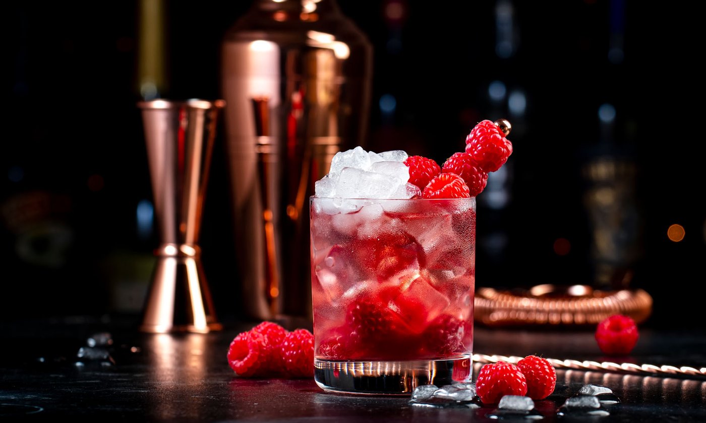 Cocktail mit Himbeeren und Crushed Ice, dahinter Shaker und Barwerkzeug in Kupfer